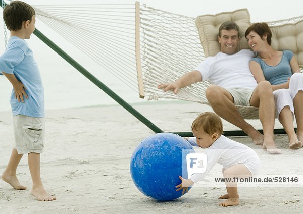 Family on beach  baby picking up ball while parents watch from hammock