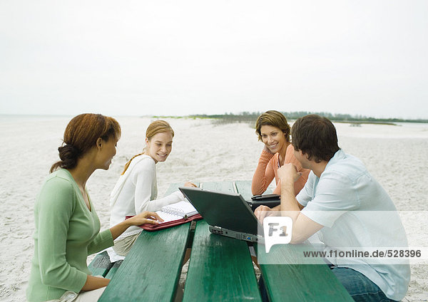 Vier lässig gekleidete Erwachsene am Picknicktisch am Strand  mit Laptop und Agenden