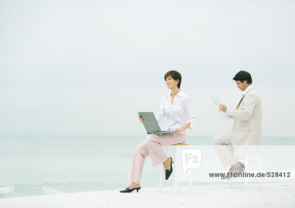 Zwei Geschäftsleute sitzen auf Hockern am Strand  einer mit Laptop  der andere mit Zeitung lesen