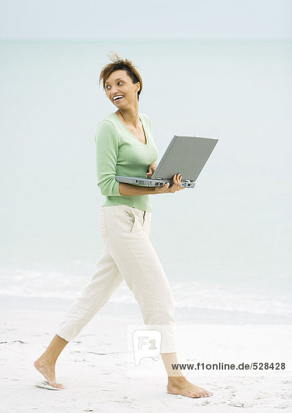 Frau geht am Strand spazieren  trägt einen offenen Laptop und schaut über die Schulter.