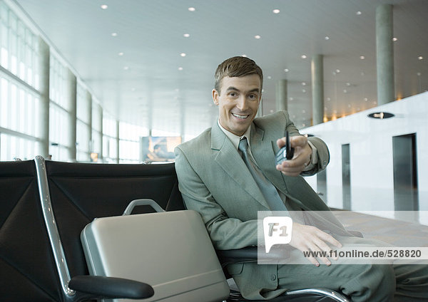 Geschäftsmann sitzt in der Flughafenlounge und hält das Handy in Richtung Kamera.