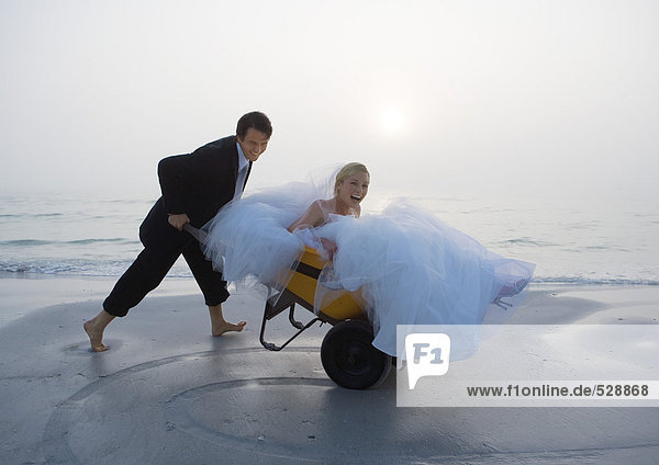 Groom pushing bride in wheelbarrow on beach