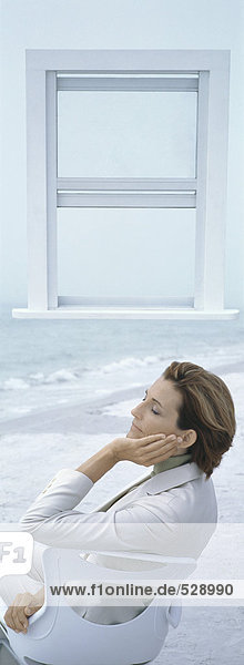 Geschäftsfrau sitzt auf einem Stuhl am Strand mit geschlossenen Augen  offenes Fenster im Hintergrund