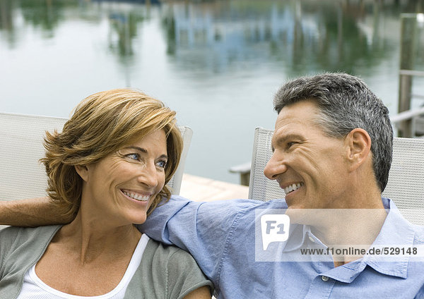 Ein reifes Paar sitzt am Wasser und lächelt sich an.