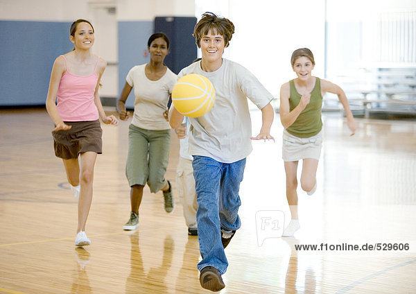 Teenager-Junge läuft mit Basketball in der Turnhalle  gefolgt von Klassenkameraden
