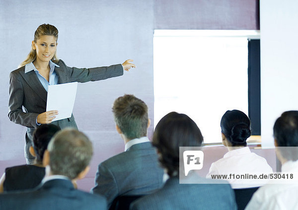Executives sitting in seminar  woman standing facing group  gesturing to screen
