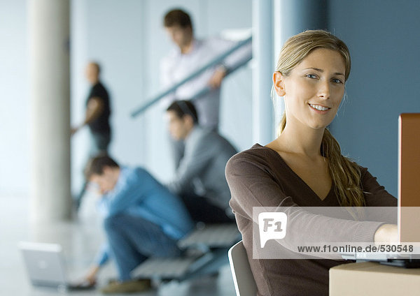Junge Frau mit Laptop  lächelnd vor der Kamera  beschäftigtes Büro im Hintergrund