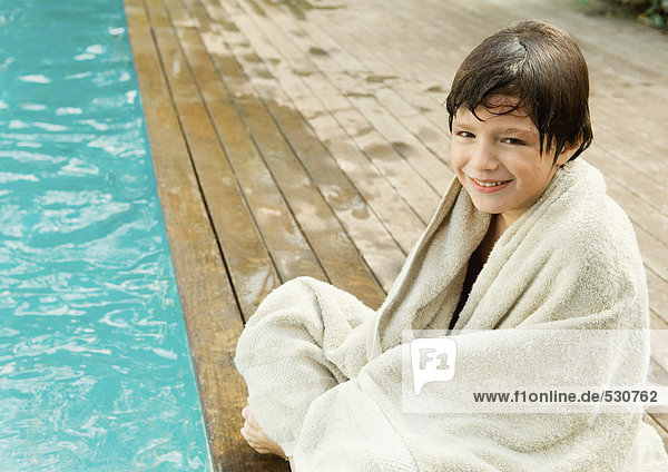 Little boy sitting by edge of swimming pool  wrapped in towel