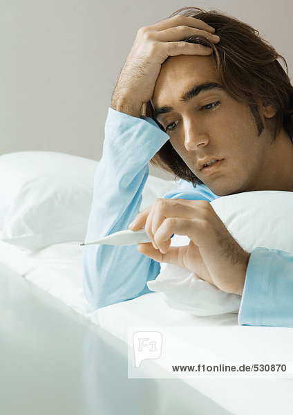 Mann im Bett liegend  auf Thermometer schauend