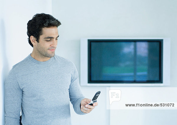 Mann steht  schaut auf Handy  Widescreen-TV an der Wand im Hintergrund