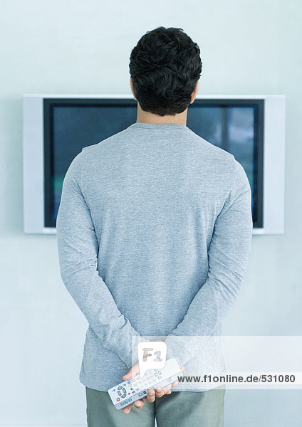 Mann steht vor dem Breitbild-Fernseher  hält zwei Fernbedienungen  Rückansicht