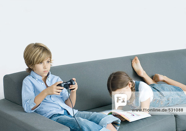 Boy sitting on sofa  holding joystick  girl next to him reading