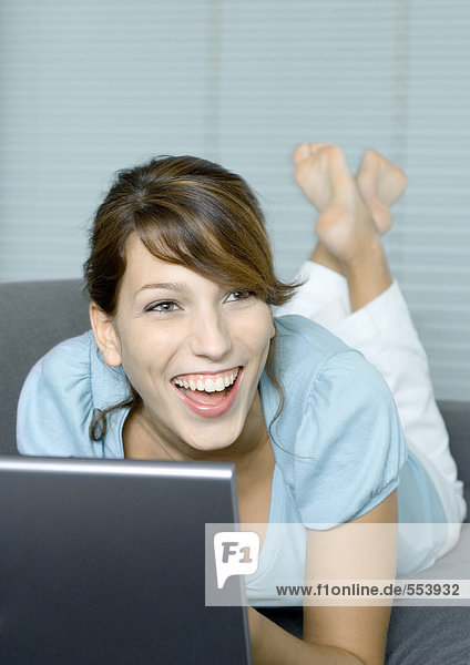 Junge Frau auf der Couch liegend  mit Laptop  lachend