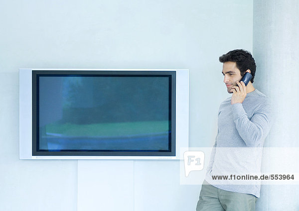 Mann  der sich mit dem Telefon an die Wand lehnt  neben dem Breitbild-Fernseher