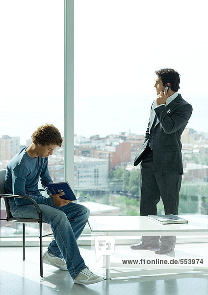 Warteraum  lässig gekleideter junger Mann sitzt auf einem Stuhl  während der Geschäftsmann Telefon am Fenster benutzt.