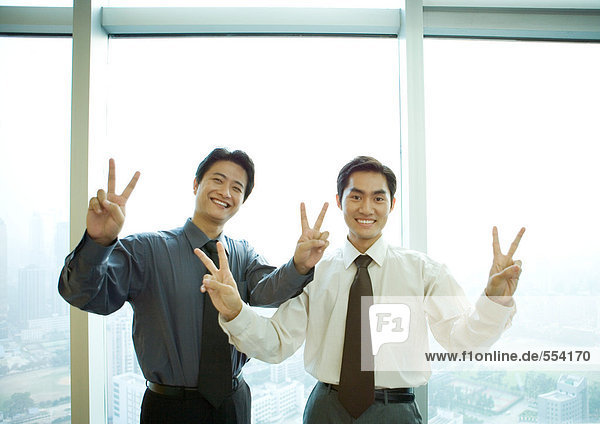 Zwei Geschäftsleute stehen am Fenster im Hochhaus  machen ein Siegeszeichen  lächeln in die Kamera.
