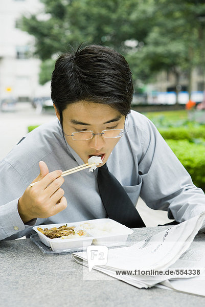 Businessman eating lunch with chopsticks and reading newspaper