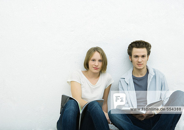 Teenager-Mädchen und junger Mann sitzen auf dem Boden mit Büchern und schauen in die Kamera.