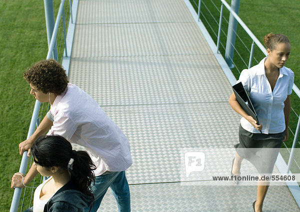 Teenage couple leaning against rail of walkway while businesswoman walks by