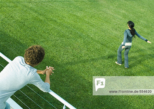 Teenager-Junge  der sich auf das Geländer stützt und sieht  wie sich ein Teenager-Mädchen auf Gras dreht.