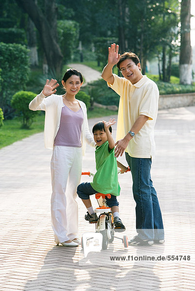 Junge auf dem Fahrrad  Eltern auf beiden Seiten  winkend vor der Kamera