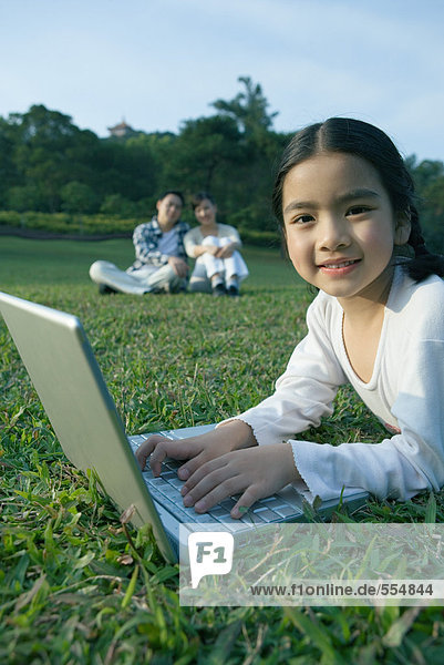Mädchen im Gras liegend mit Laptop  Eltern im Gras sitzend im Hintergrund