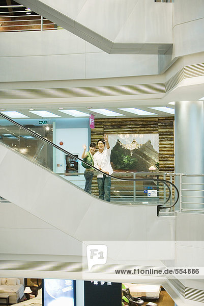 Paar nimmt Rolltreppe im Einkaufszentrum  winkt vor der Kamera  in mittlerer Entfernung