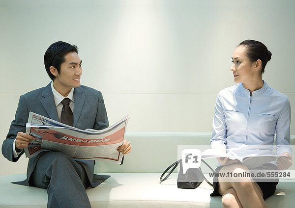 Businessman and woman sitting on waiting room bench  smiling at each other