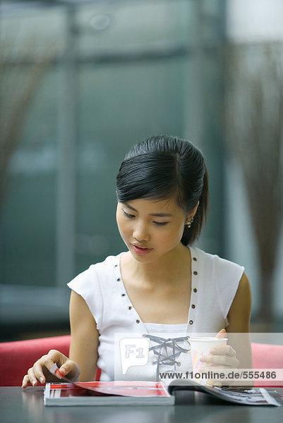 Junge Frau sitzt am Tisch  schaut auf die Zeitschrift  hält einen Drink.