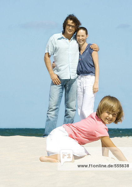 Mädchen graben im Sand  während die Eltern im Hintergrund stehen.
