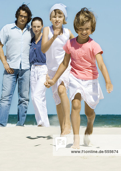 Mädchen laufen am Strand  während die Eltern im Hintergrund stehen.