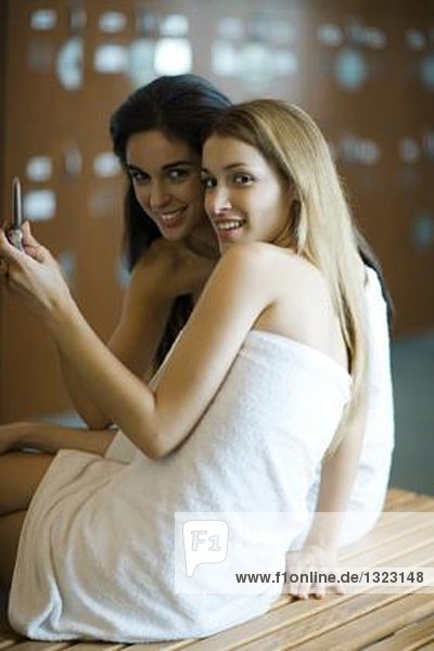 Junge Frauen sitzen in Handtüchern im Umkleideraum und fotografieren mit dem Handy.
