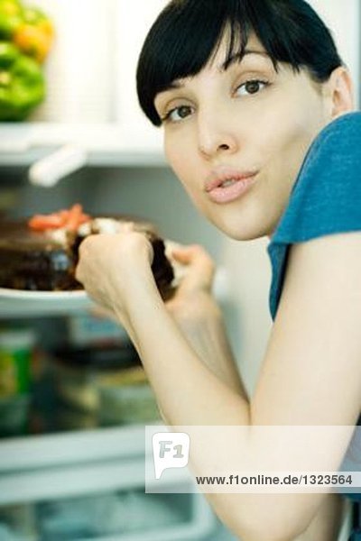 Junge Frau nimmt ein Stück Kuchen aus dem Kühlschrank  schaut über die Schulter in die Kamera