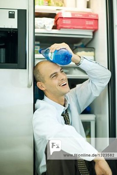Mann sitzt neben geöffneter Kühlschranktür und hält eine Flasche Wasser gegen die Stirn.