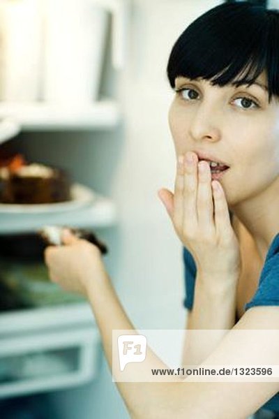 Junge Frau nimmt ein Stück Kuchen aus dem Kühlschrank  schaut in die Kamera  bedeckt den Mund