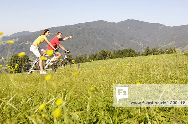 Young couple riding mountain bike in meadow  side view  mountains in background