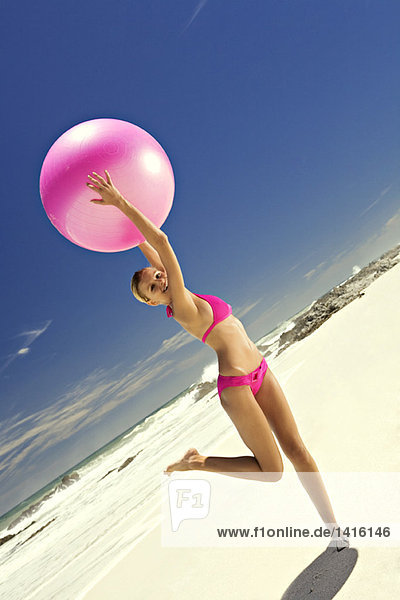 Junge lächelnde Frau im rosa Bikini am Strand  die einen großen Ball hebt.