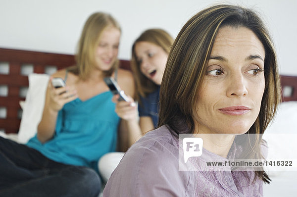 2 Teenager-Mädchen sitzen auf dem Bett  benutzen Mobiltelefone  denkende Frau im Vordergrund