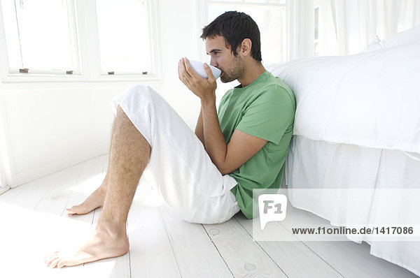 Young man drinking coffee  leaning against bed