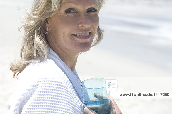 Porträt einer lächelnden Frau am Strand,  die ein Glas Wasser hält.