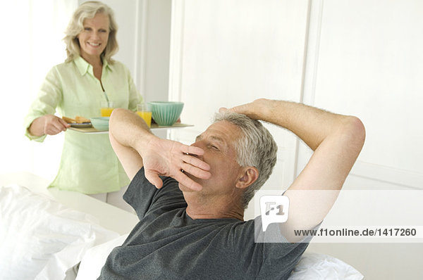 Woman bringing breakfast to man yawning in bed
