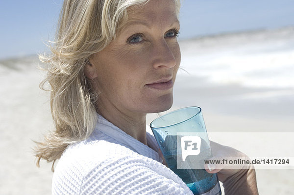 Porträt einer denkenden Frau am Strand  die ein Glas Wasser hält.