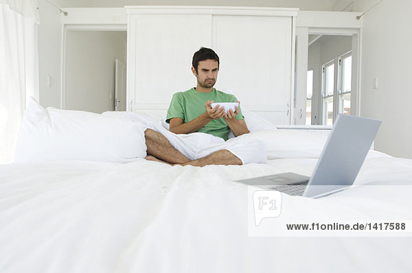 Junger Mann sitzt im Schneidersitz auf einem Bett und schaut auf den Laptop.