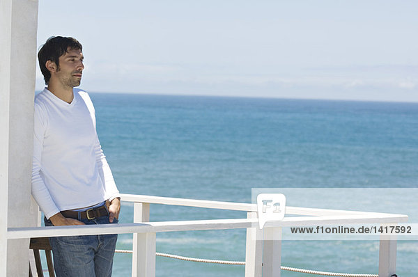 Junger denkender Mann  der sich auf einer Terrasse an die Wand lehnt  Meer im Hintergrund