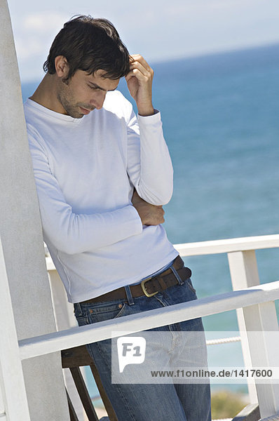 Junger denkender Mann  der sich auf einer Terrasse an die Wand lehnt  Meer im Hintergrund