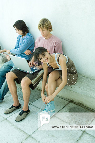 Gruppe junger Freunde auf der Bank sitzend  eine junge Frau bindet Schuhbänder und lächelt in die Kamera.