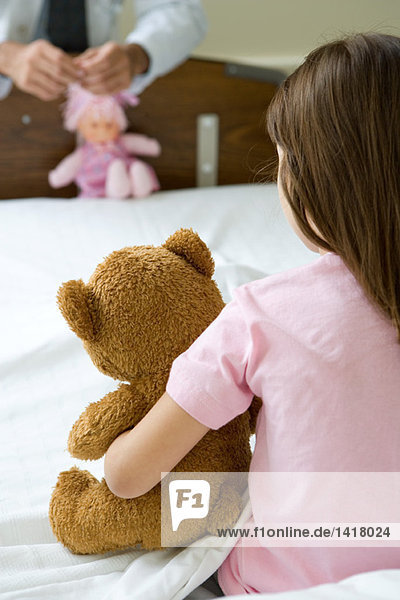 Mädchen im Krankenhausbett sitzend  Teddybär haltend  Ärztin im Gesicht  Puppe haltend