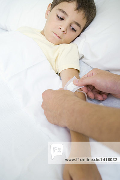 Junge liegt im Krankenhausbett und beobachtet Mann legt Verband über Infusionsschlauch