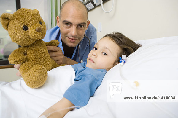 Mädchen im Krankenhausbett liegend  männlicher Assistenzarzt neben ihr haltend Teddybär