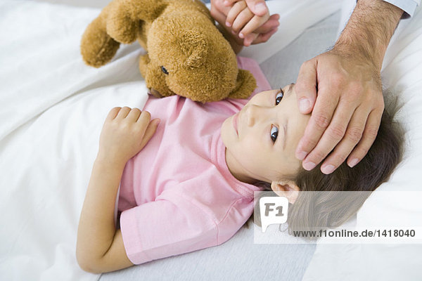 Mädchen im Bett liegend mit Teddybär  Mann hält ihre Hand auf die andere Hand auf die Stirn gelegt